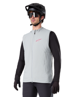 Gilet thermique Alpinestars A-Dura, gris titane, gilet de cyclisme isolant, porté sur un haut noir à manches longues avec un casque de VTT et des gants noirs, arborant un logo rose sur la poitrine et des poches latérales zippées verticales
