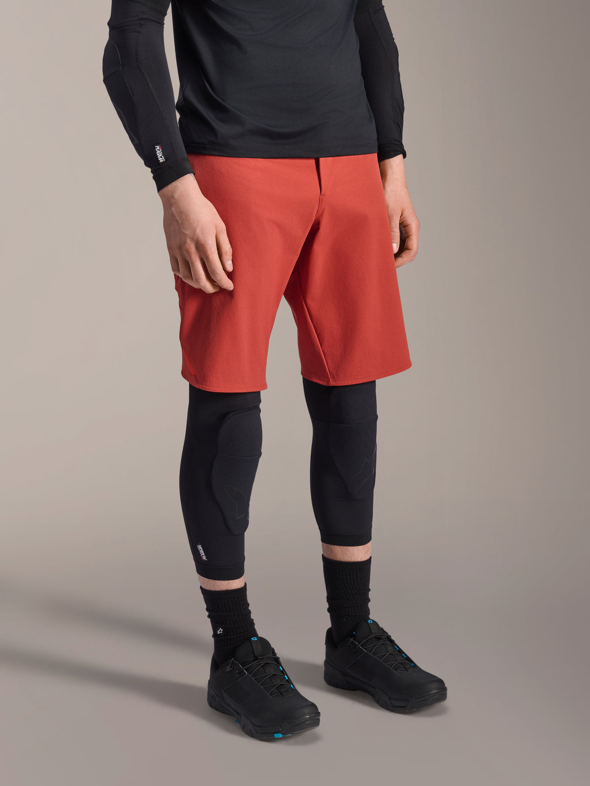 Alpinestars A-Motion Plasma Knee Sleeve, genouillère de VTT, noir, porté avec un short rouge et des chaussures de VTT noires, design à enfiler discret avec protections profilées et maille respirante.