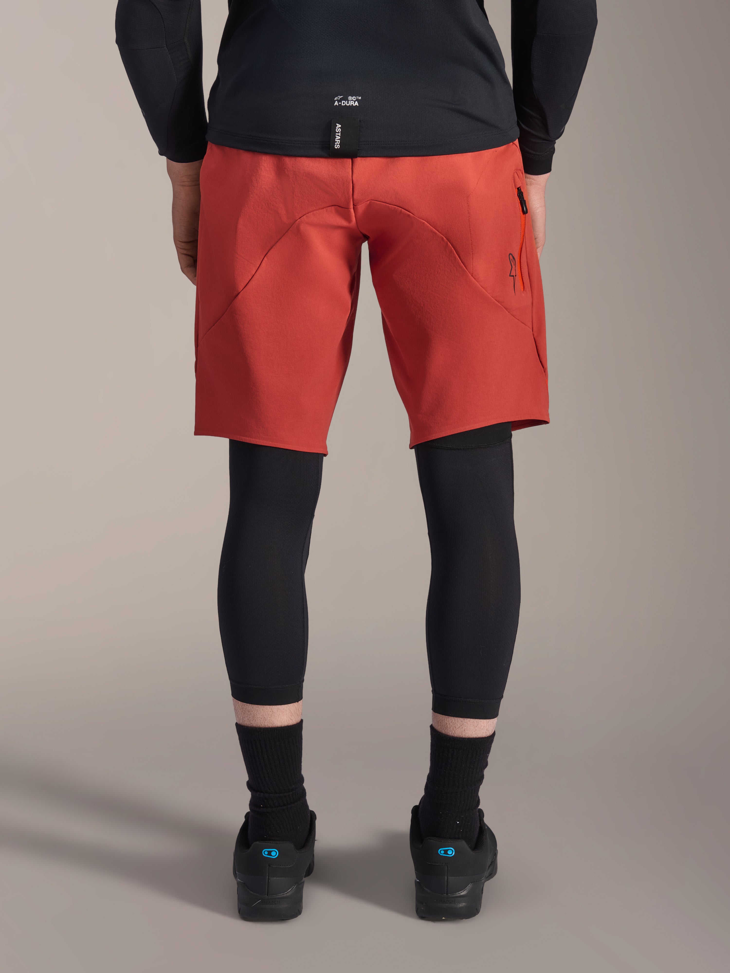 Alpinestars A-Motion Plasma Knee Sleeve, protections de genou VTT noires, vue de dos sous un short de VTT rouge avec un maillot noir et des chaussures de cyclisme techniques, équipement de protection pour le vélo tout-terrain