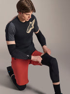 Manchon de genou Alpinestars A-Motion Plasma, protection de genou VTT, noir, porté par un cycliste en position agenouillée, associé à un t-shirt noir et gris avec un logo doré, un short rouge et des coudières noires, équipement de protection professionnel pour VTT