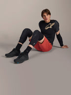 Alpinestars A-Motion Plasma Knee Sleeve, genouillères de VTT noires, portées par un mannequin homme assis en tenue de cyclisme complète avec un short rouge et un tee-shirt technique noir, mettant en avant le design de protection discret.
