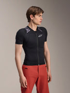 Male model wearing Alpinestars A-Motion Plasma Protection Jacket, Short Sleeve, Black Camo with gray camouflage shoulder panels, featuring integrated shoulder and chest protectors, white star logo on chest, breathable mesh fabric, paired with red pants