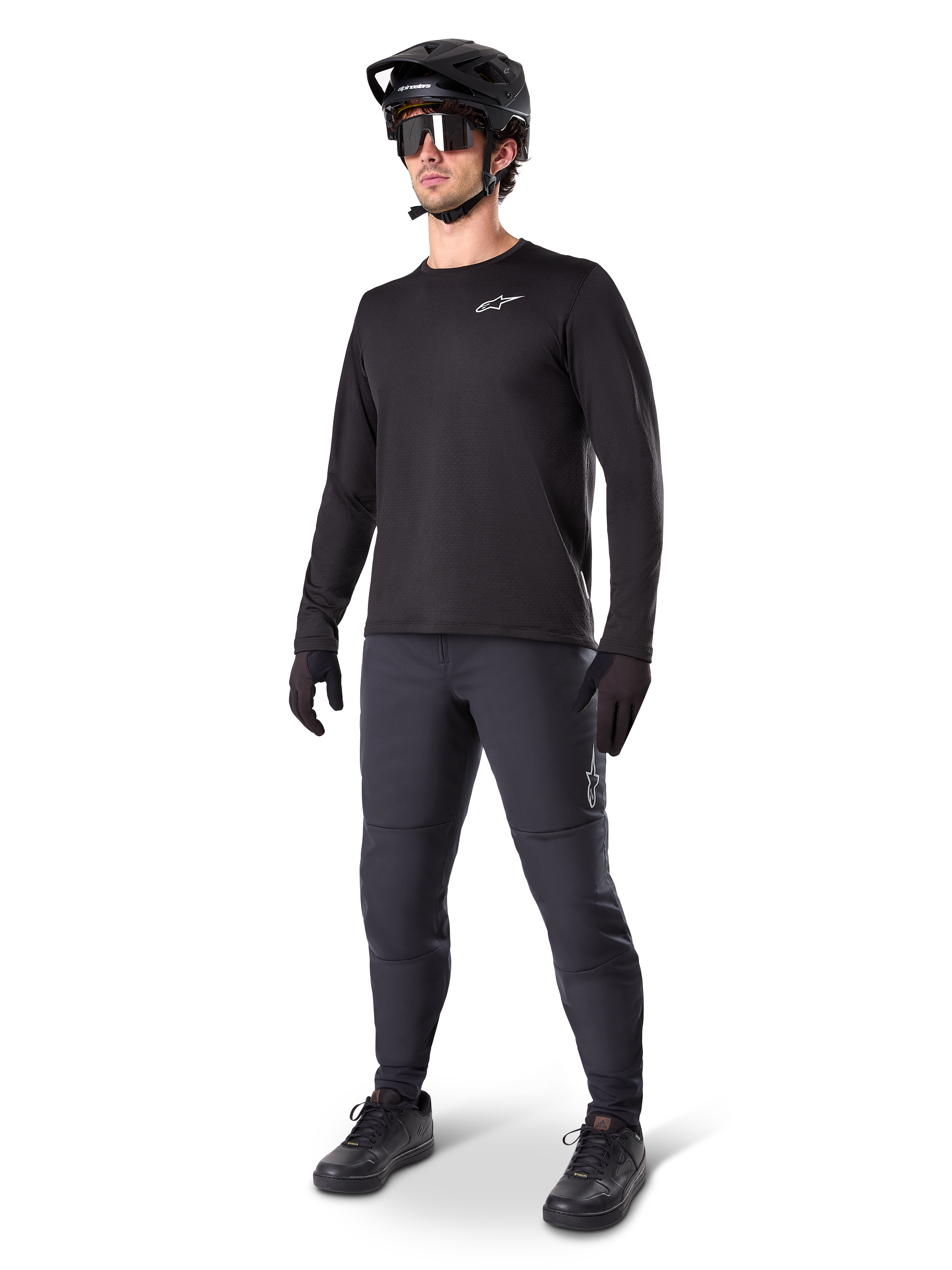 Cycliste de VTT portant le pantalon thermique Alpinestars A-Dura noir, avec un maillot à manches longues noir, un casque noir et des lunettes de soleil, logo Alpinestars argenté sur la cuisse gauche, conçu pour le trail par temps froid.