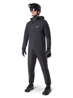 Vététiste portant le pantalon imperméable Alpinestars A-Dura noir, associé à une veste imperméable à capuche noire assortie, un casque de VTT noir avec visière, des lunettes de cyclisme teintées et des gants noirs, équipement professionnel complet pour le trail par tous les temps