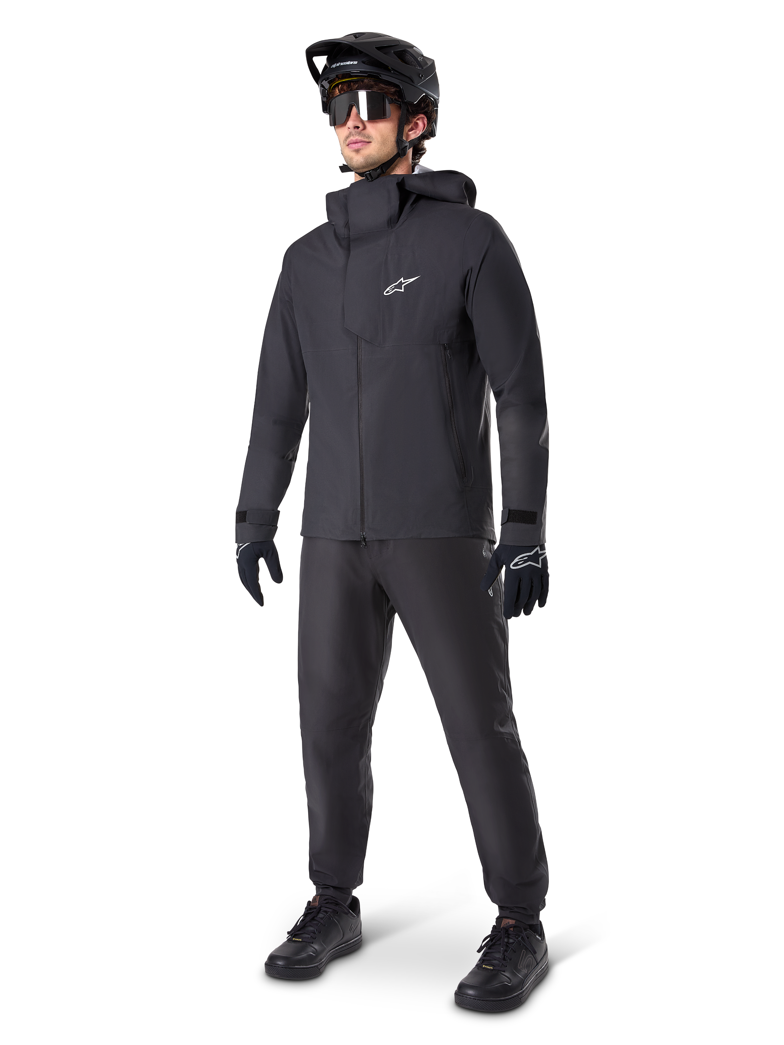 Vététiste portant le pantalon imperméable Alpinestars A-Dura noir, associé à une veste imperméable à capuche noire assortie, un casque de VTT noir avec visière, des lunettes de cyclisme teintées et des gants noirs, équipement professionnel complet pour le trail par tous les temps