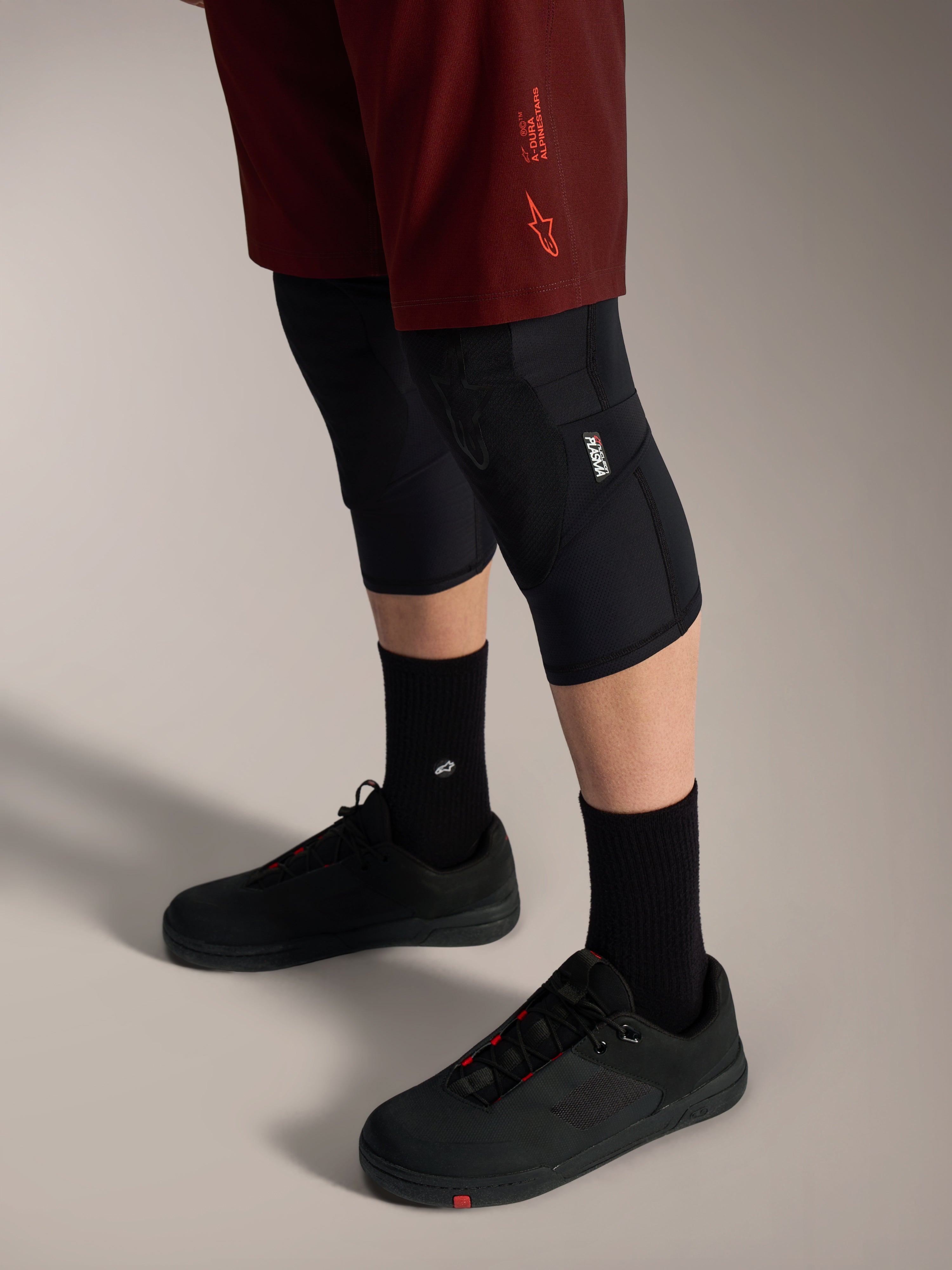 Shorts Alpinestars A-Dura Ride bordeaux, coupe extensible et logo signature, portés avec genouillères et chaussures noires.