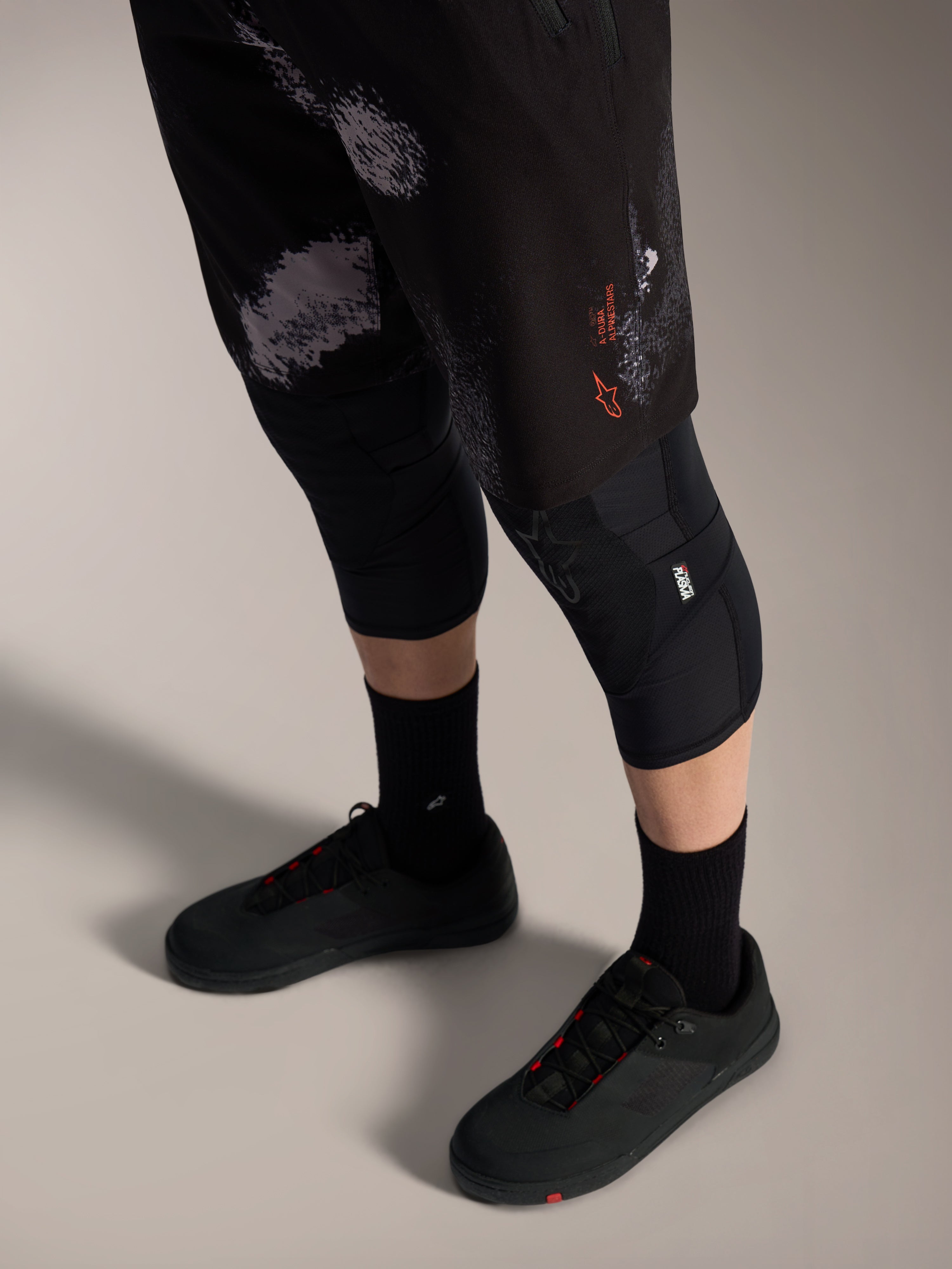 Shorts Alpinestars A-Dura Ride Black Camo, équipement VTT avec logo orange, genouillères et chaussures de vélo.