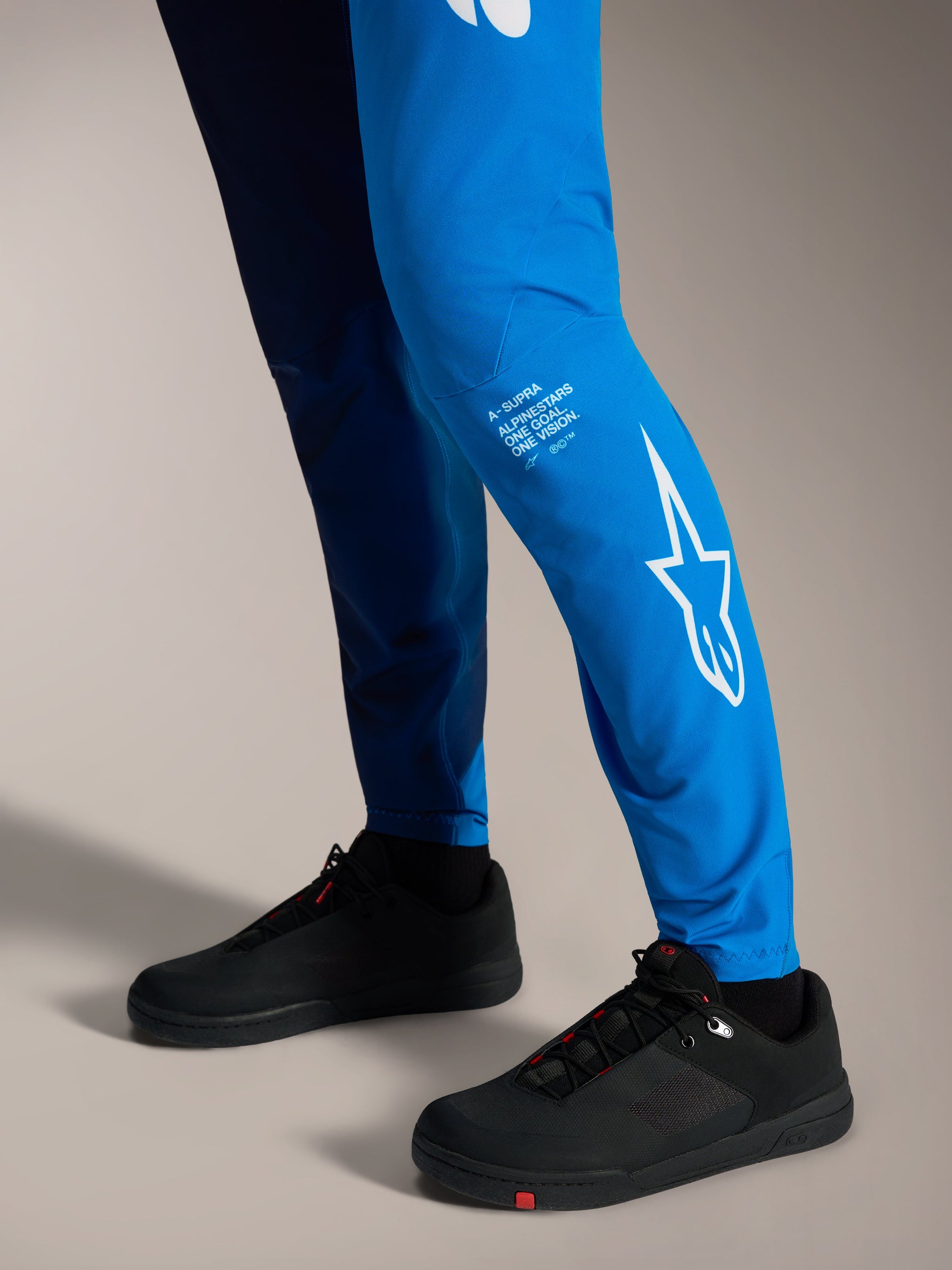Pantalons A-Supra Elite bleu vif délavé, logo Astars blanc sur le bas de la jambe et chaussures de VTT noires.