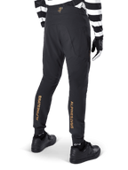 Alpinestars A-Aria Elite Pants, Black MTB Pants, rear view, featuring vertical gold Alpinestars branding on the back of the legs, worn with a black and white striped jersey and black shoes, performance cycling apparel for mountain biking