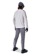 Alpinestars A-Aria Elite Pants, MTB Cycling Pants, Dark Gray, rear view of a rider wearing a full outfit, featuring white Alpinestars branding on the lower legs, performance fit with laser perforations, paired with a light gray long-sleeve jersey and gray MTB helmet
