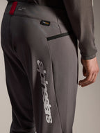 Pantalon Alpinestars A-Dura Elite, pantalon de VTT gris foncé, vue rapprochée de la hanche et de la cuisse, avec grand logo Alpinestars vertical blanc, étiquette Cordura, poche zippée et détail rouge à la taille