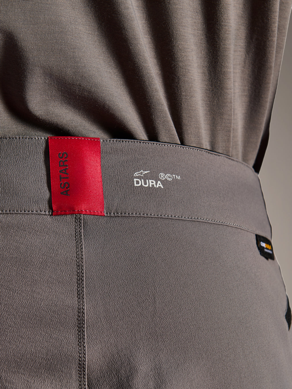 Pantalon Alpinestars A-Dura Elite, pantalon de VTT, gris foncé, gros plan sur la ceinture montrant une étiquette ASTARS rouge et un logo DURA blanc, doté d'un tissu Cordura texturé et de détails de marquage premium.
