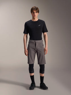 Alpinestars A-Dura Elite Shorts, short de VTT gris foncé, porté avec un t-shirt noir à col rond et des genouillères noires, vêtement de cyclisme de performance à hauteur de genou avec fermeture à pression à l'avant et tissu Cordura durable.