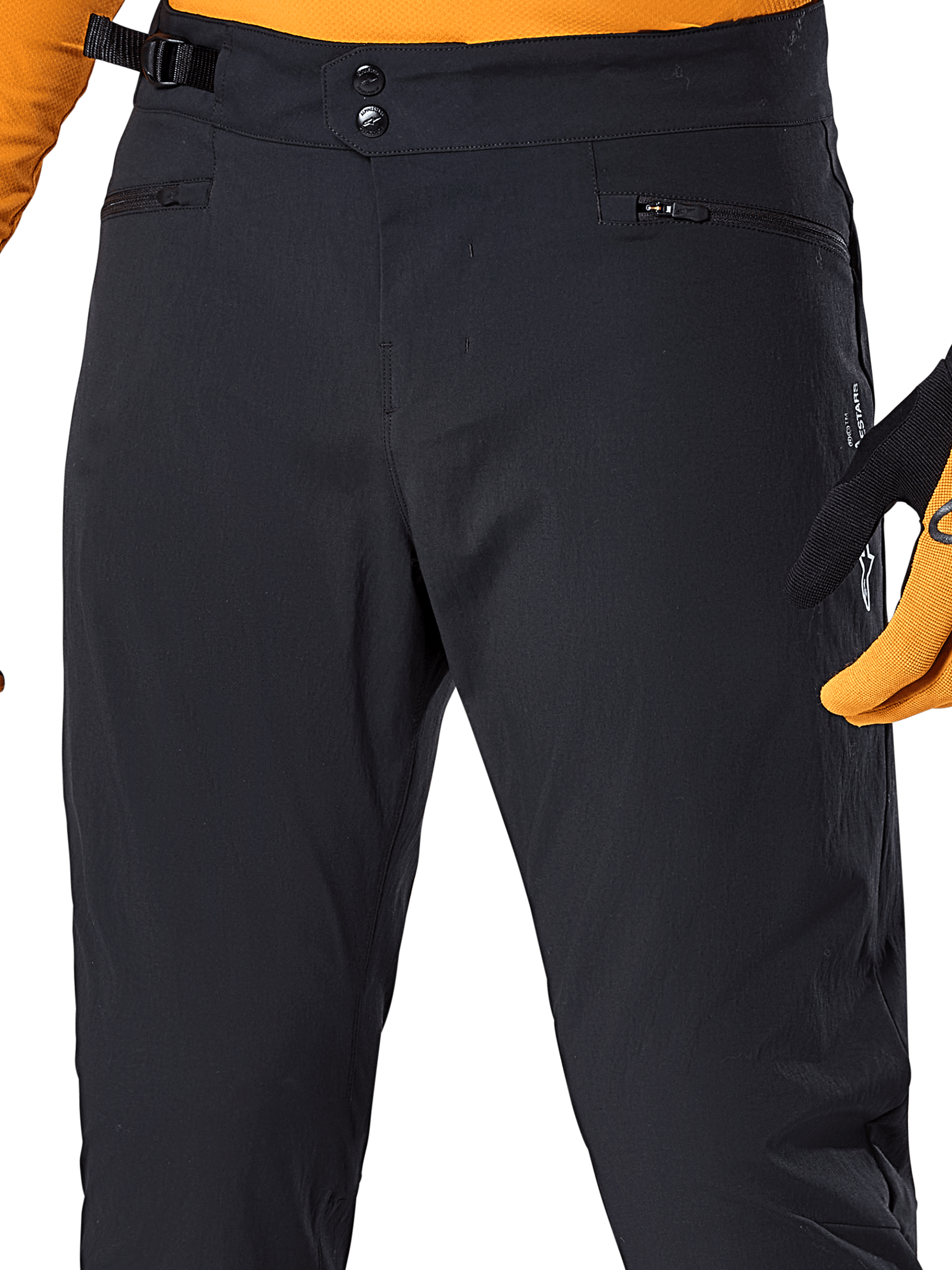 Pantalon Alpinestars A-Dura, pantalon de cyclisme VTT noir, vue rapprochée présentant la double fermeture à pression à la taille, l'ajusteur latéral réglable et les poches zippées, porté avec un maillot et des gants orange