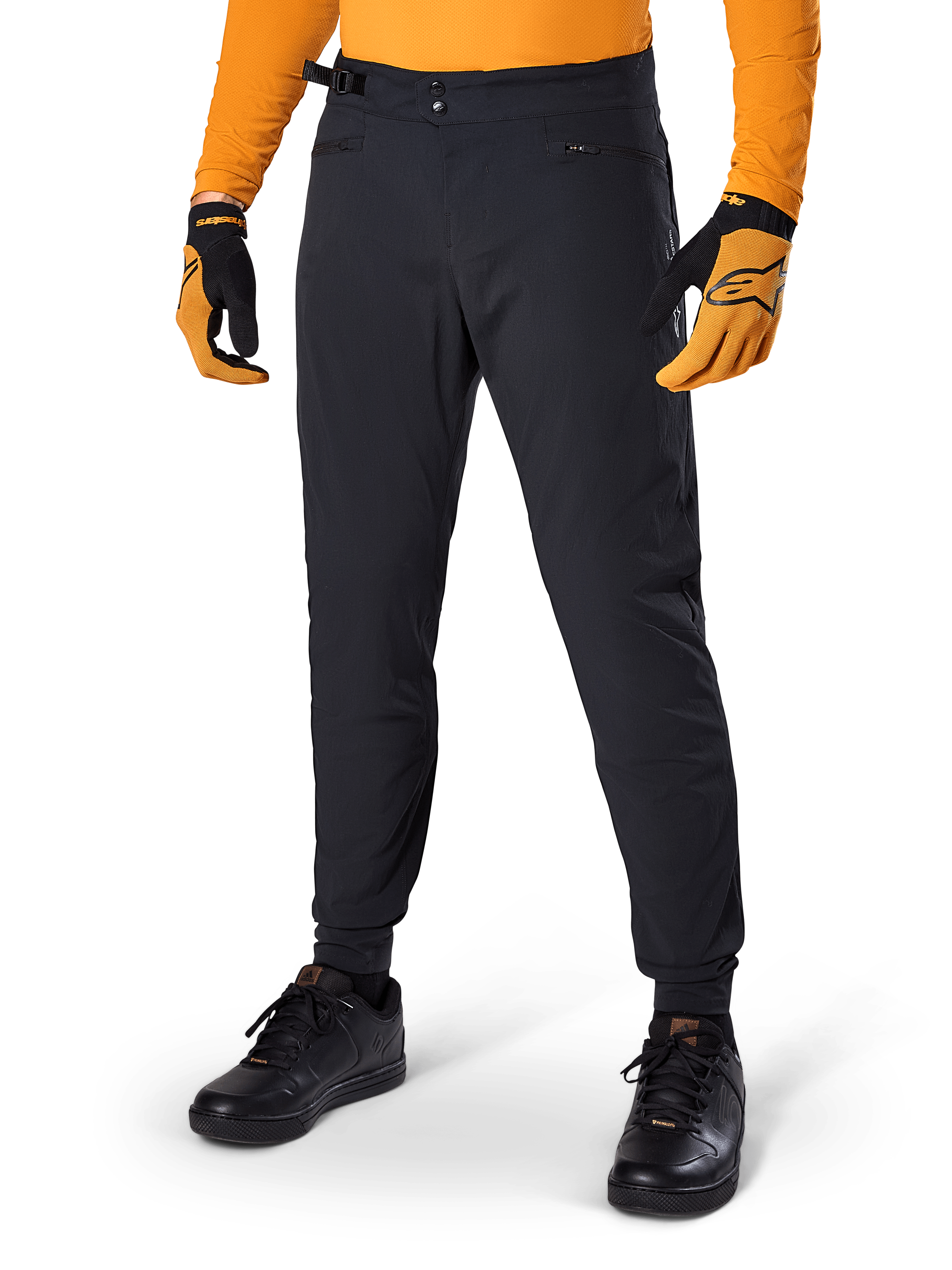 Alpinestars A-Dura Pants, pantalon de VTT, noir, serrages latéraux à la taille et poches zippées, porté avec un maillot et des gants orange, coupe régulière sportive pour la pratique du VTT