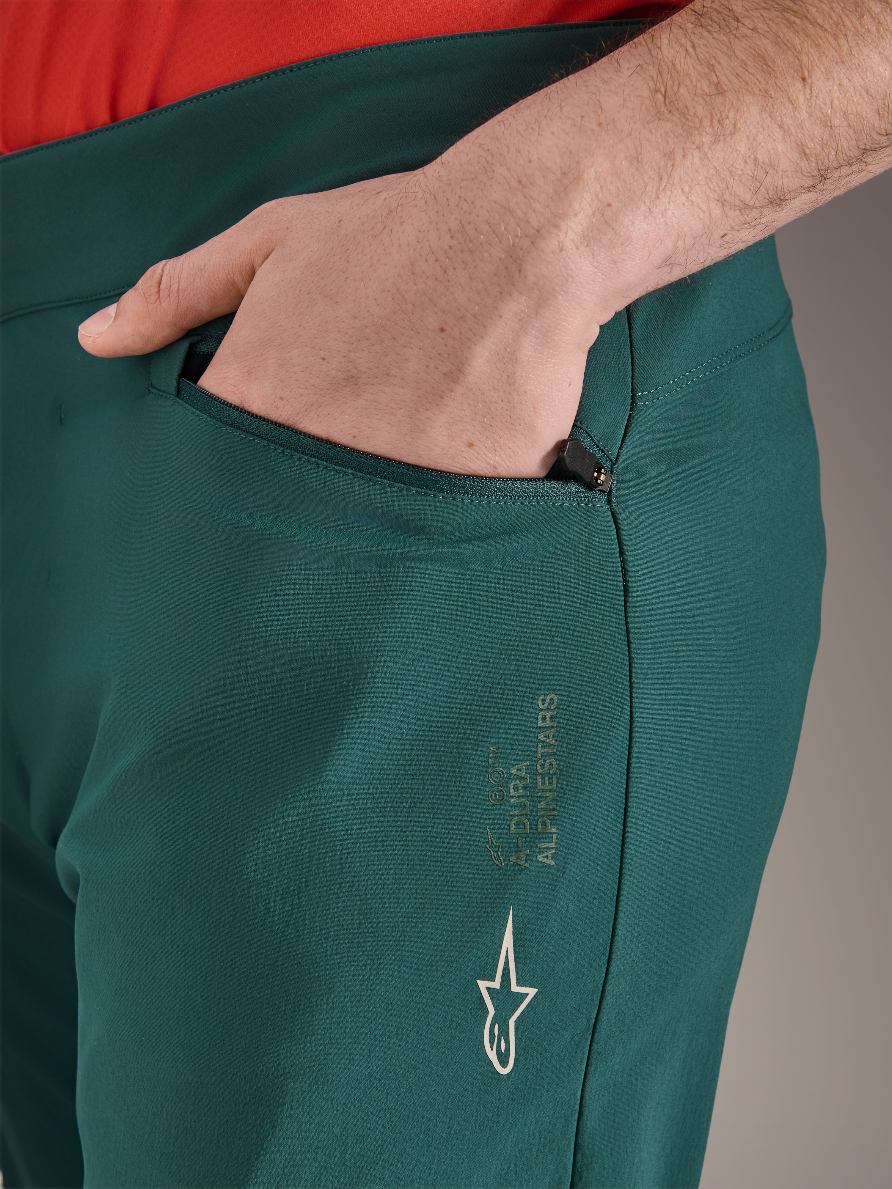 Pantalon Alpinestars A-Dura vert Dark Pine, gros plan d'une poche zippée avec une main, logo Alpinestars blanc et marquage sur la cuisse, pantalon de VTT durable