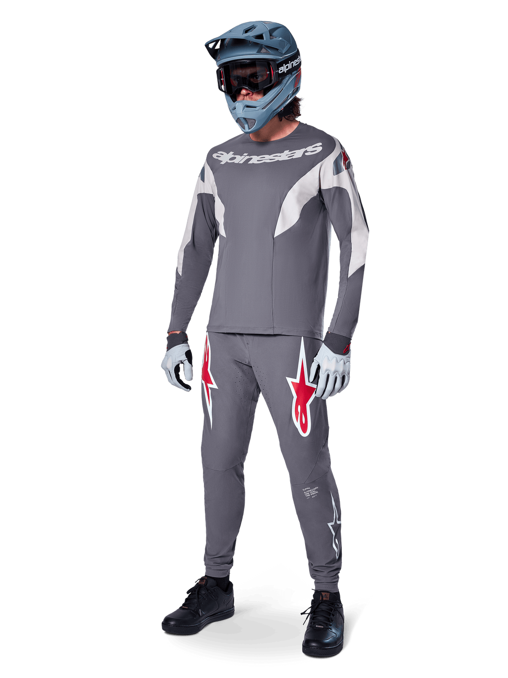 Pantalon Alpinestars A-Supra Race en gris foncé, avec des logos rouges et blancs sur les cuisses, porté par un pilote avec un maillot gris assorti, un casque intégral bleu-gris et des gants gris clair, équipement haute performance pour la course VTT