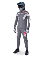 Pantalon Alpinestars A-Supra Race en gris foncé, avec des logos rouges et blancs sur les cuisses, porté par un pilote avec un maillot gris assorti, un casque intégral bleu-gris et des gants gris clair, équipement haute performance pour la course VTT