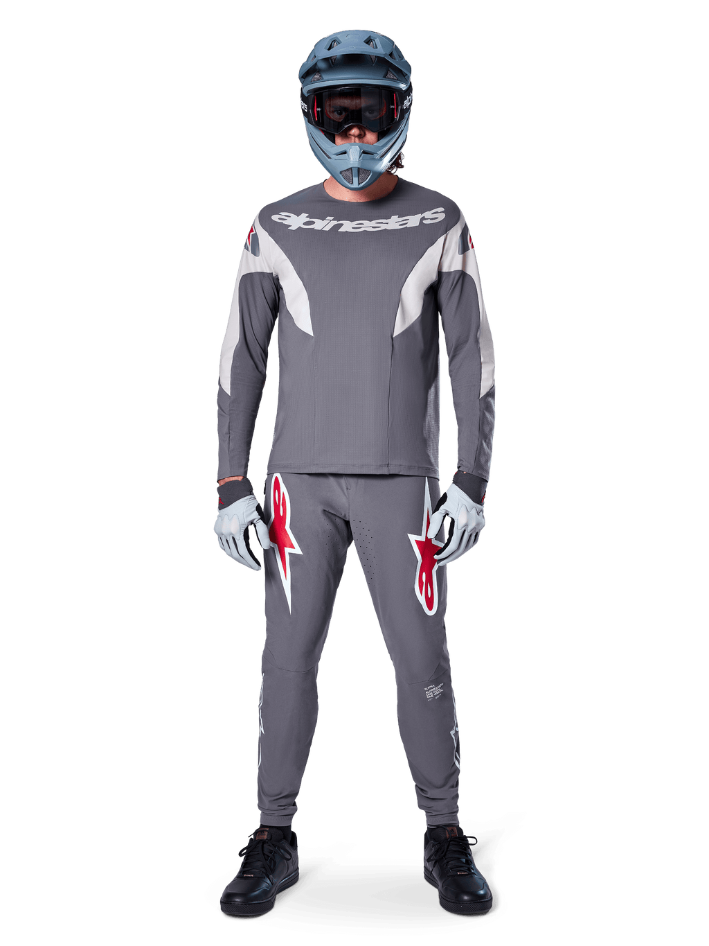 Pilote de VTT portant le pantalon de course Alpinestars A-Supra gris foncé, avec des logos rouges vifs sur les cuisses, un maillot gris assorti, un casque intégral bleu et des chaussures noires pour un équipement complet de course tout-terrain