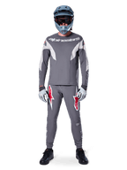 Pilote de VTT portant le pantalon de course Alpinestars A-Supra gris foncé, avec des logos rouges vifs sur les cuisses, un maillot gris assorti, un casque intégral bleu et des chaussures noires pour un équipement complet de course tout-terrain