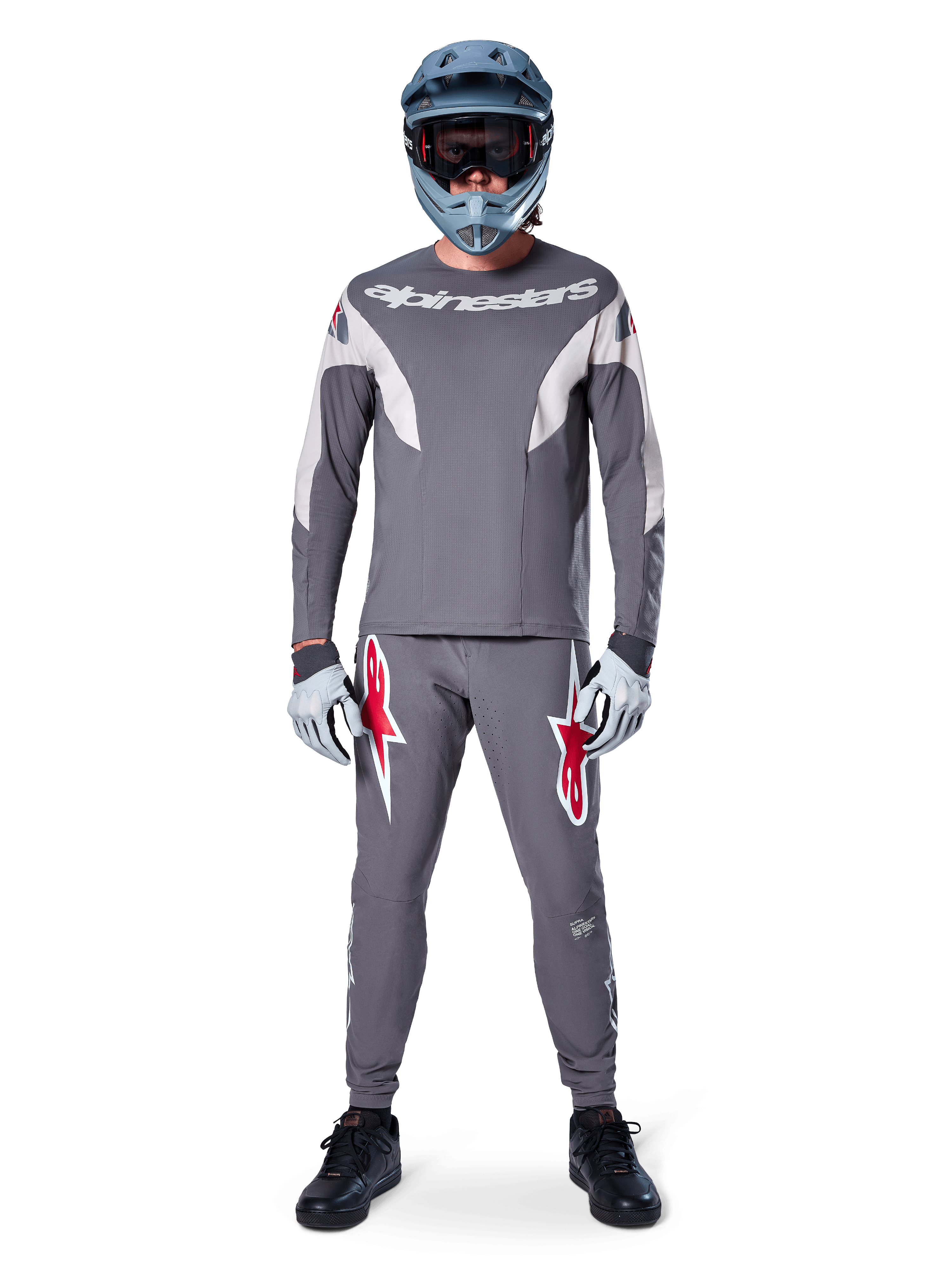 Pilote de VTT portant le pantalon de course Alpinestars A-Supra gris foncé, avec des logos rouges vifs sur les cuisses, un maillot gris assorti, un casque intégral bleu et des chaussures noires pour un équipement complet de course tout-terrain