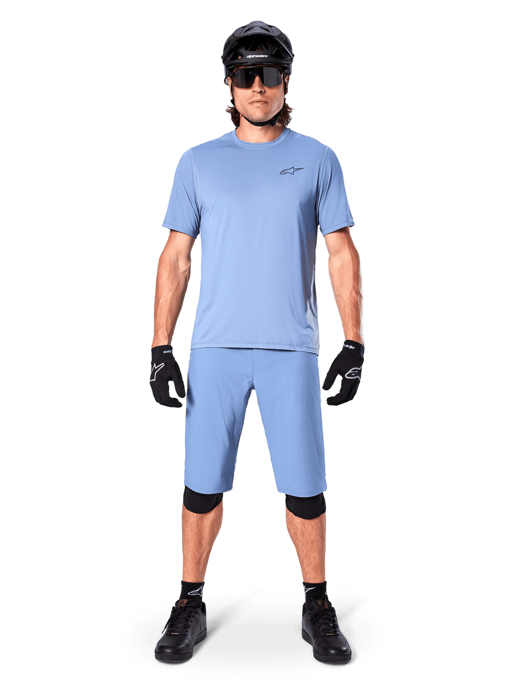 Model wearing Alpinestars A-Dura Shorts in Infinity Blue, matching light blue jersey, black mountain bike helmet, black sunglasses, black gloves and knee pads, complete MTB outfit for trail riding