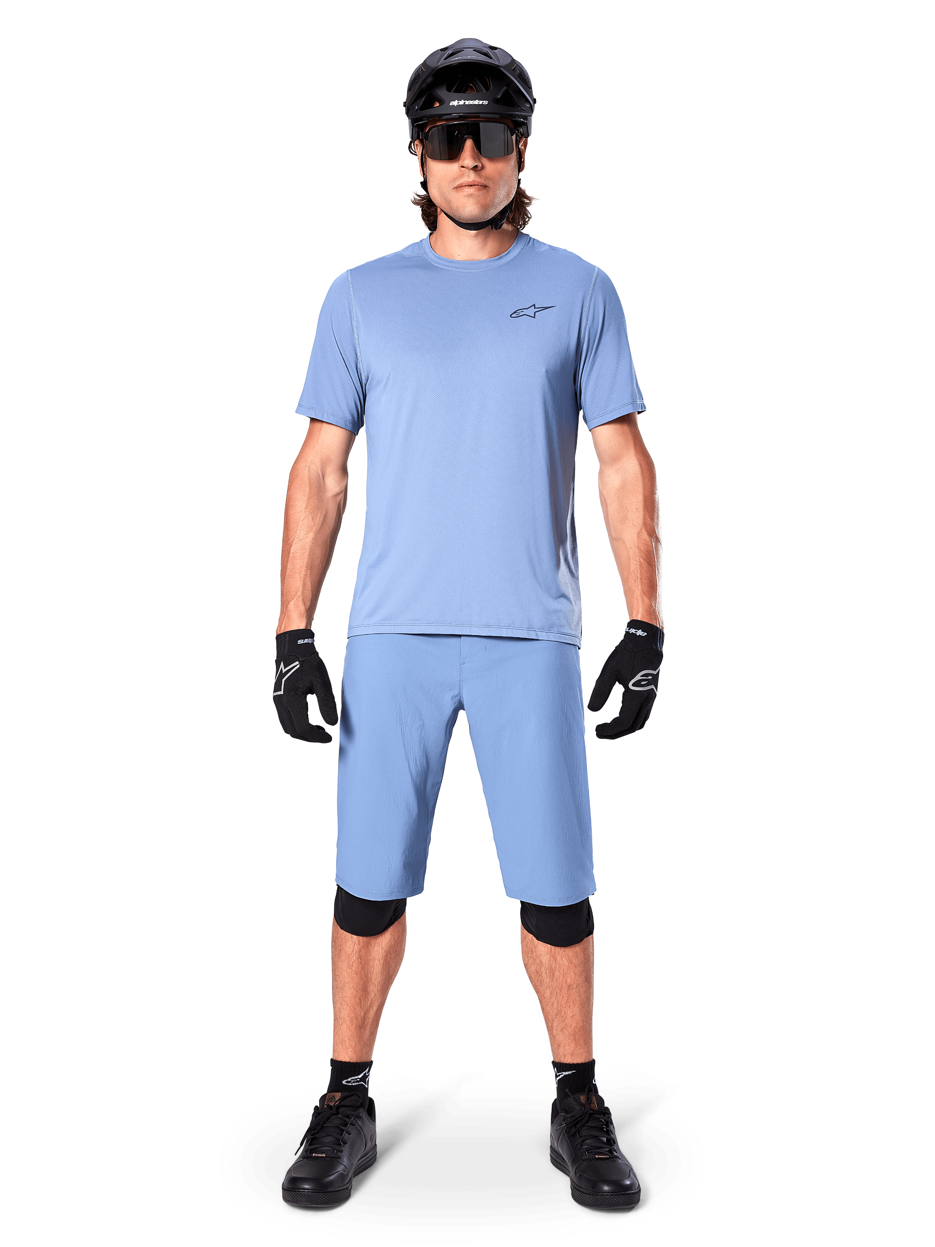 Model wearing Alpinestars A-Dura Shorts in Infinity Blue, matching light blue jersey, black mountain bike helmet, black sunglasses, black gloves and knee pads, complete MTB outfit for trail riding