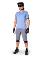 Short Alpinestars A-Dura, short de VTT, gris foncé, porté par un rider avec un maillot bleu clair, un casque de VTT noir, des gants noirs et des genouillères, tenue de VTT complète pour le trail et le tout-terrain