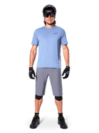 Cycliste portant un short Alpinestars A-Dura gris foncé, avec un t-shirt à manches courtes bleu clair, un casque noir, des lunettes de soleil et des genouillères noires, tenue de VTT complète pour la performance tout-terrain.