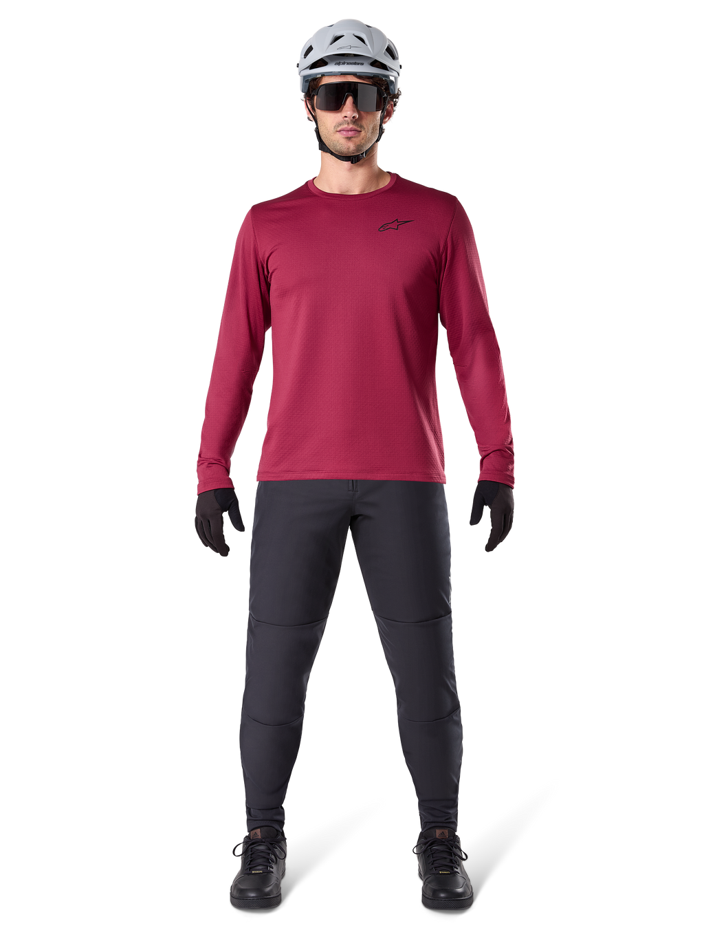 Modèle masculin portant le maillot thermique Alpinestars A-Dura bordeaux, haut de cyclisme isolé à manches longues, associé à un pantalon de VTT gris foncé, un casque blanc, des lunettes de soleil noires et des gants noirs, équipement professionnel de VTT pour temps froid