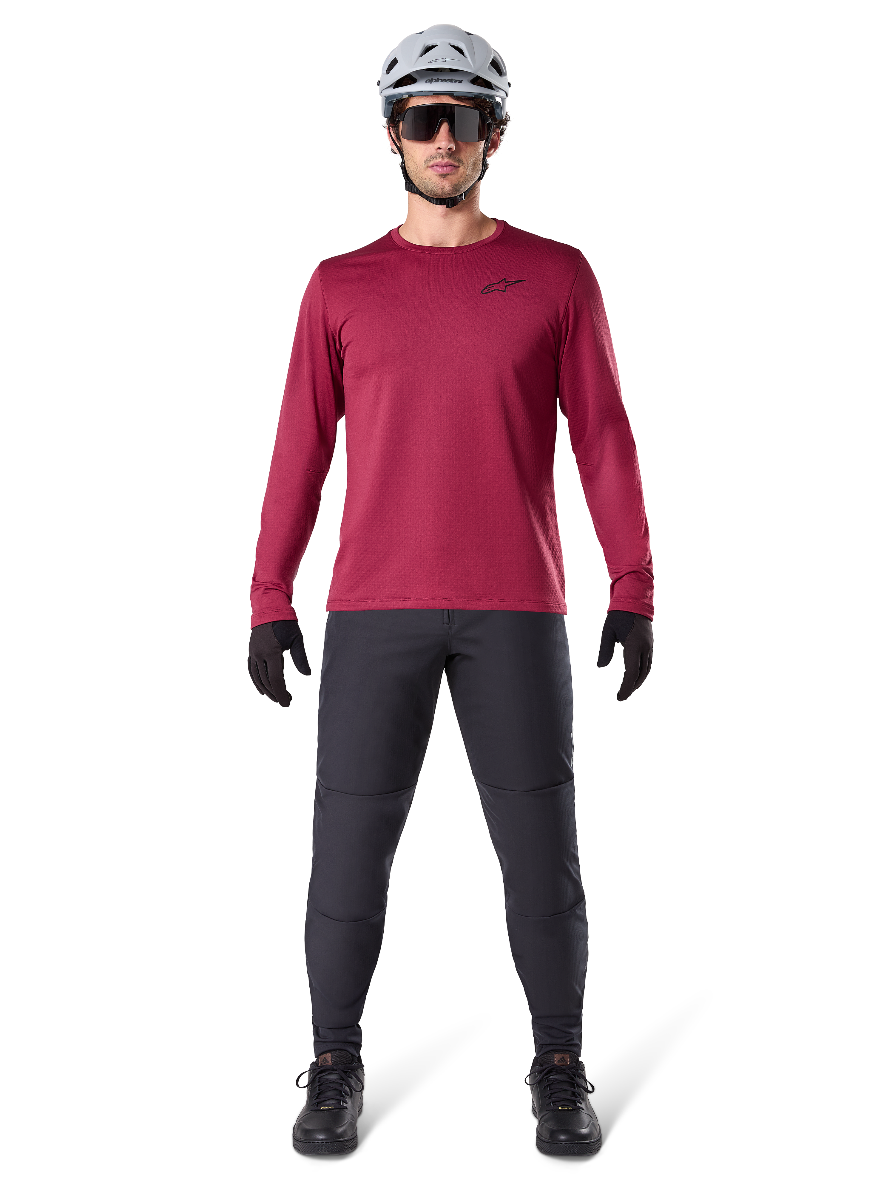Modèle masculin portant le maillot thermique Alpinestars A-Dura bordeaux, haut de cyclisme isolé à manches longues, associé à un pantalon de VTT gris foncé, un casque blanc, des lunettes de soleil noires et des gants noirs, équipement professionnel de VTT pour temps froid