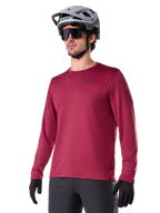 Maillot thermique Alpinestars A-Dura, maillot de VTT à manches longues, rouge bordeaux, avec doublure en polaire isolante et petit logo noir sur la poitrine, porté avec un casque gris, des lunettes de soleil noires et des gants noirs, conçu pour le cyclisme par temps froid