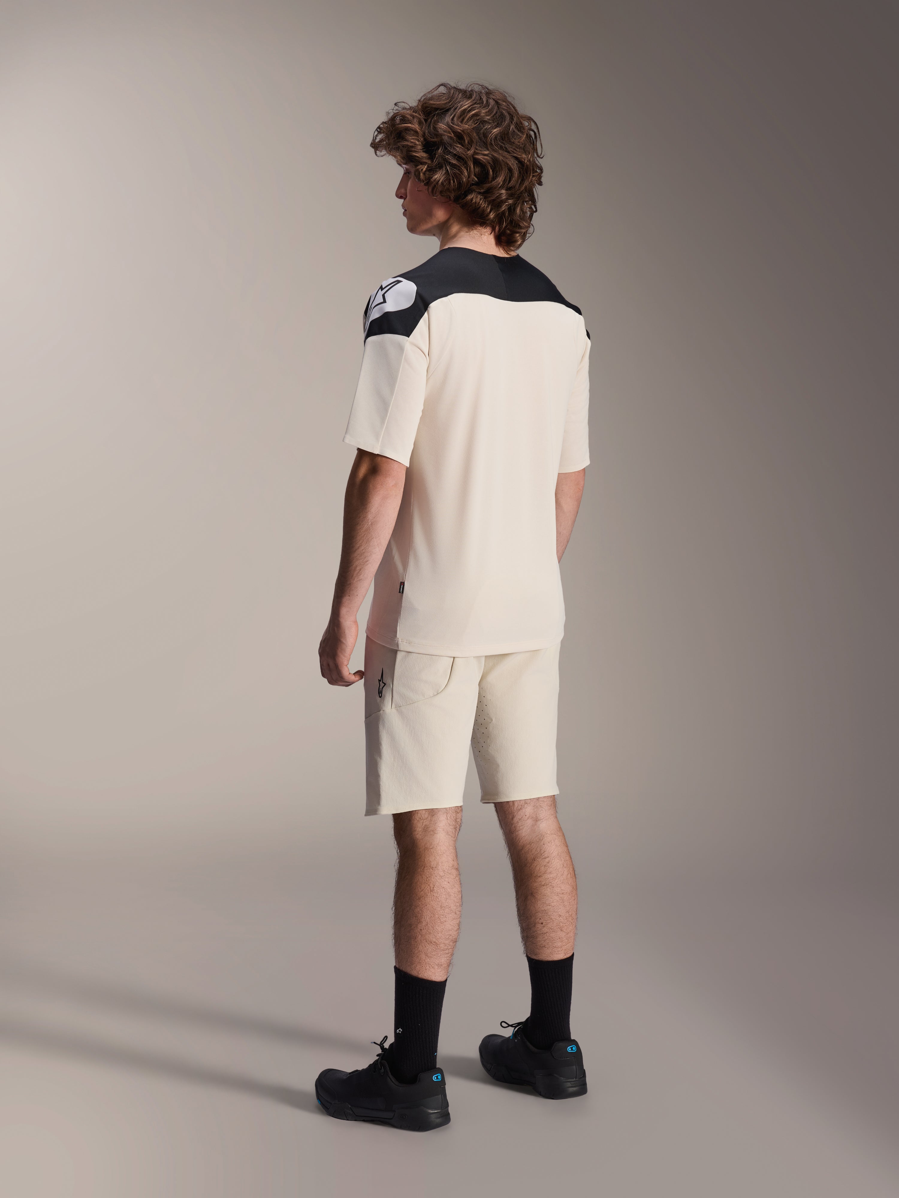 Mannequin portant le maillot à manches courtes Alpinestars A-Aria Polartec Elite en coloris Écru, vu de dos, avec des empiècements noirs aux épaules et un logo blanc, associé à un short de VTT écru assorti et des chaussures noires