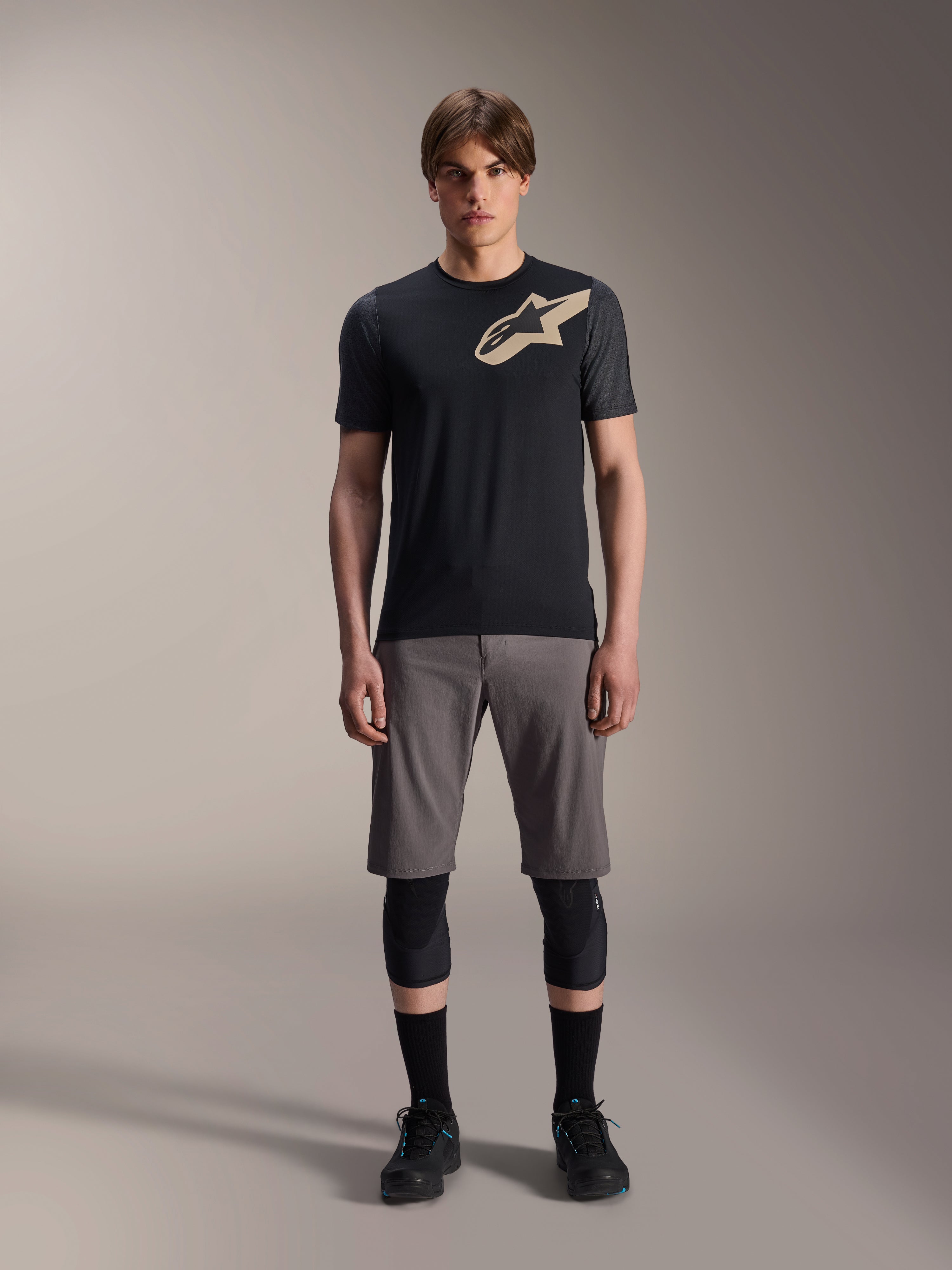 Alpinestars A-Dura Lipan Short Sleeve Jersey, Black with grey marl sleeves and tan Astars chest logo, worn by a male model with grey mountain bike shorts and black knee guards, performance MTB riding gear.
