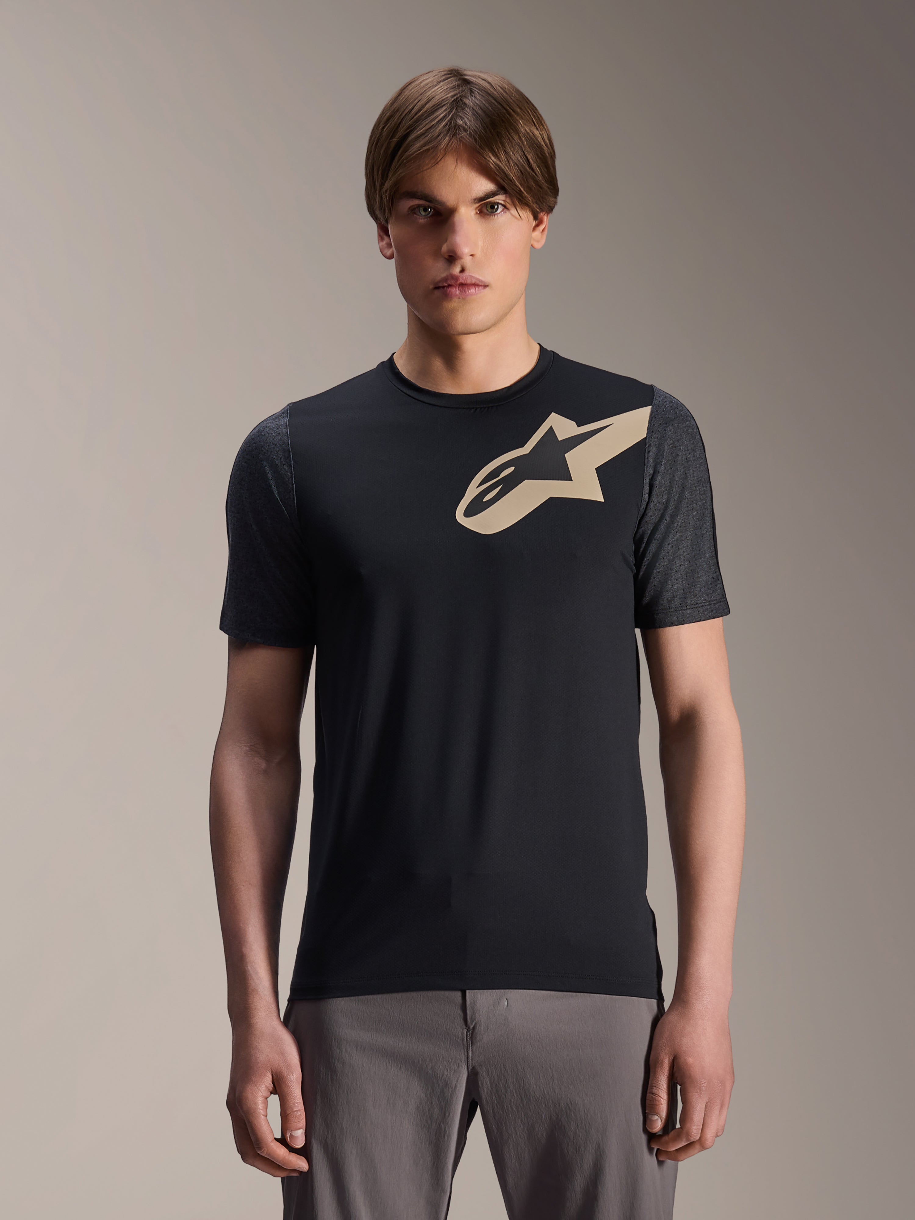 Male model wearing Alpinestars A-Dura Lipan Short Sleeve Jersey, Black MTB mountain bike jersey with beige star logo and dark gray sleeves, paired with gray cycling pants, casual performance apparel