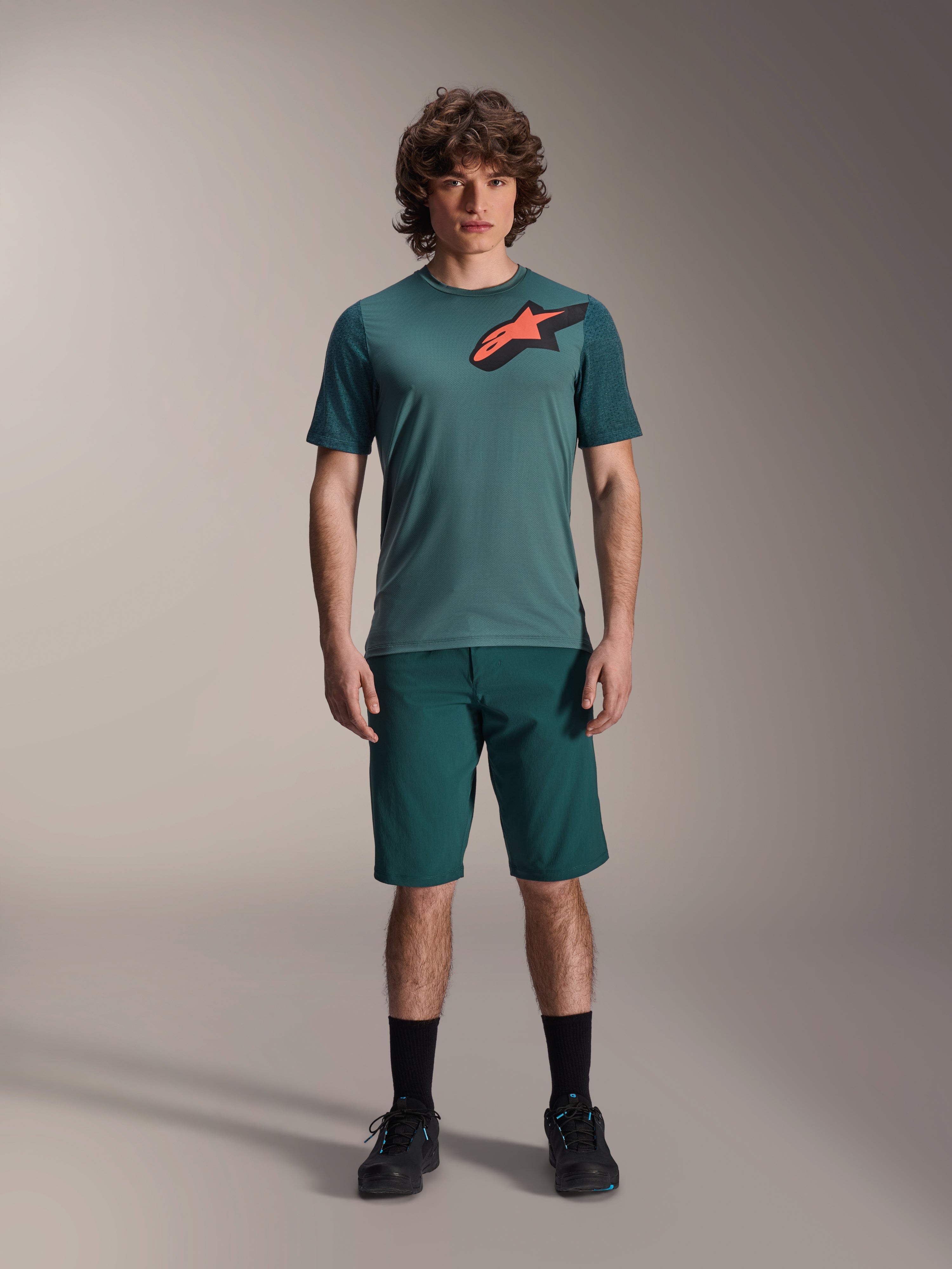 Short A-Dura Alpinestars en Dark Pine, short de VTT pour homme, présenté sur un mannequin avec un maillot à manches courtes bleu sarcelle assorti doté d'un logo étoile rouge et noir, chaussettes noires et chaussures de cyclisme, tenue de VTT décontractée et sportive