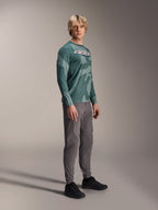 Alpinestars A-Dura Hollow Camo Long Sleeve Jersey, Silver Pine Camo, worn by a model with grey pants and black shoes, featuring a green camouflage pattern and a bold white ASTARS logo on the chest, high-performance MTB apparel