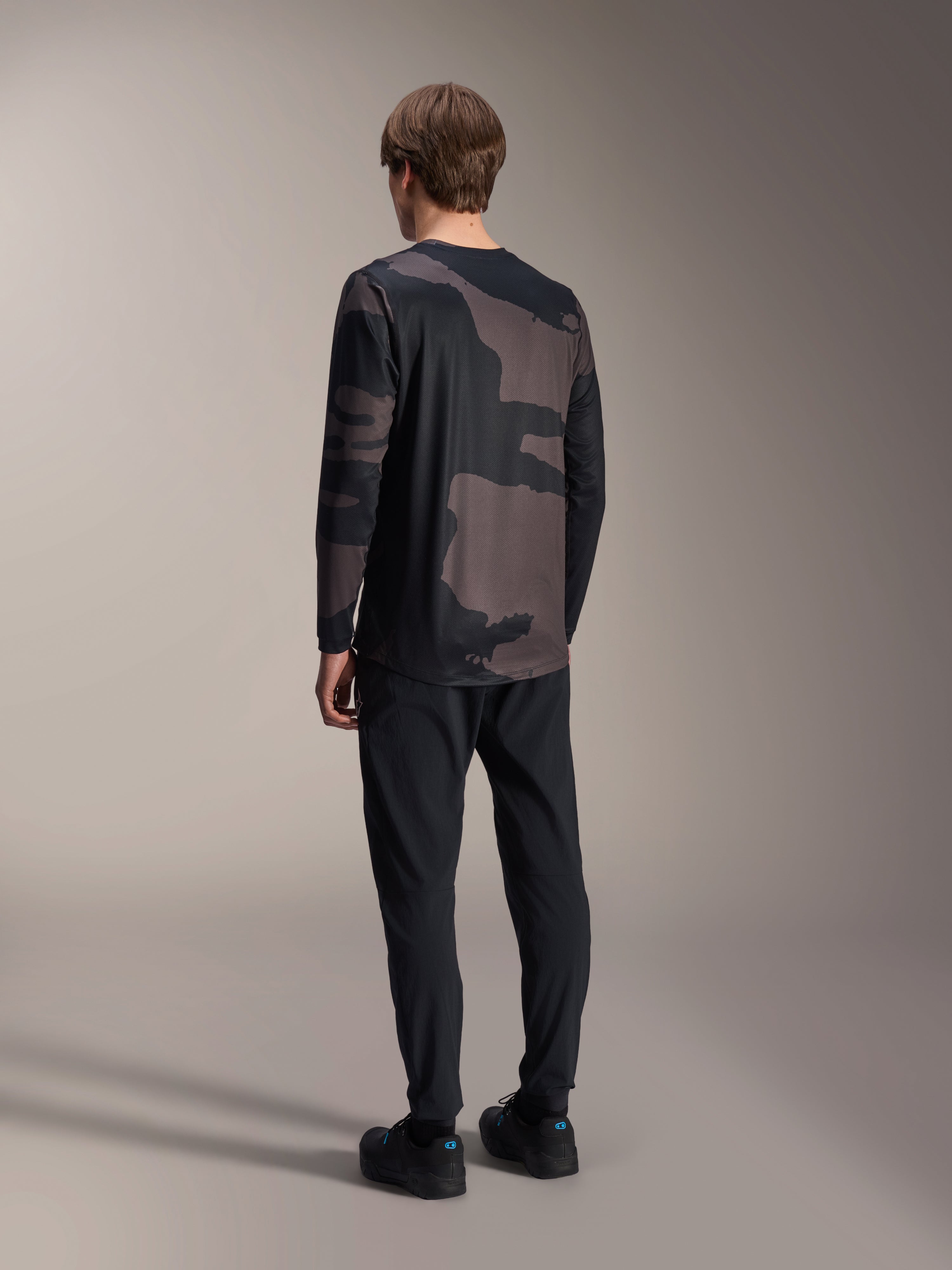 Alpinestars A-Dura Hollow Camo Long Sleeve Jersey, Black Camo, rear view of model, technical mountain bike jersey with dropped tail hem, paired with black pants and cycling shoes, casual off-road performance gear