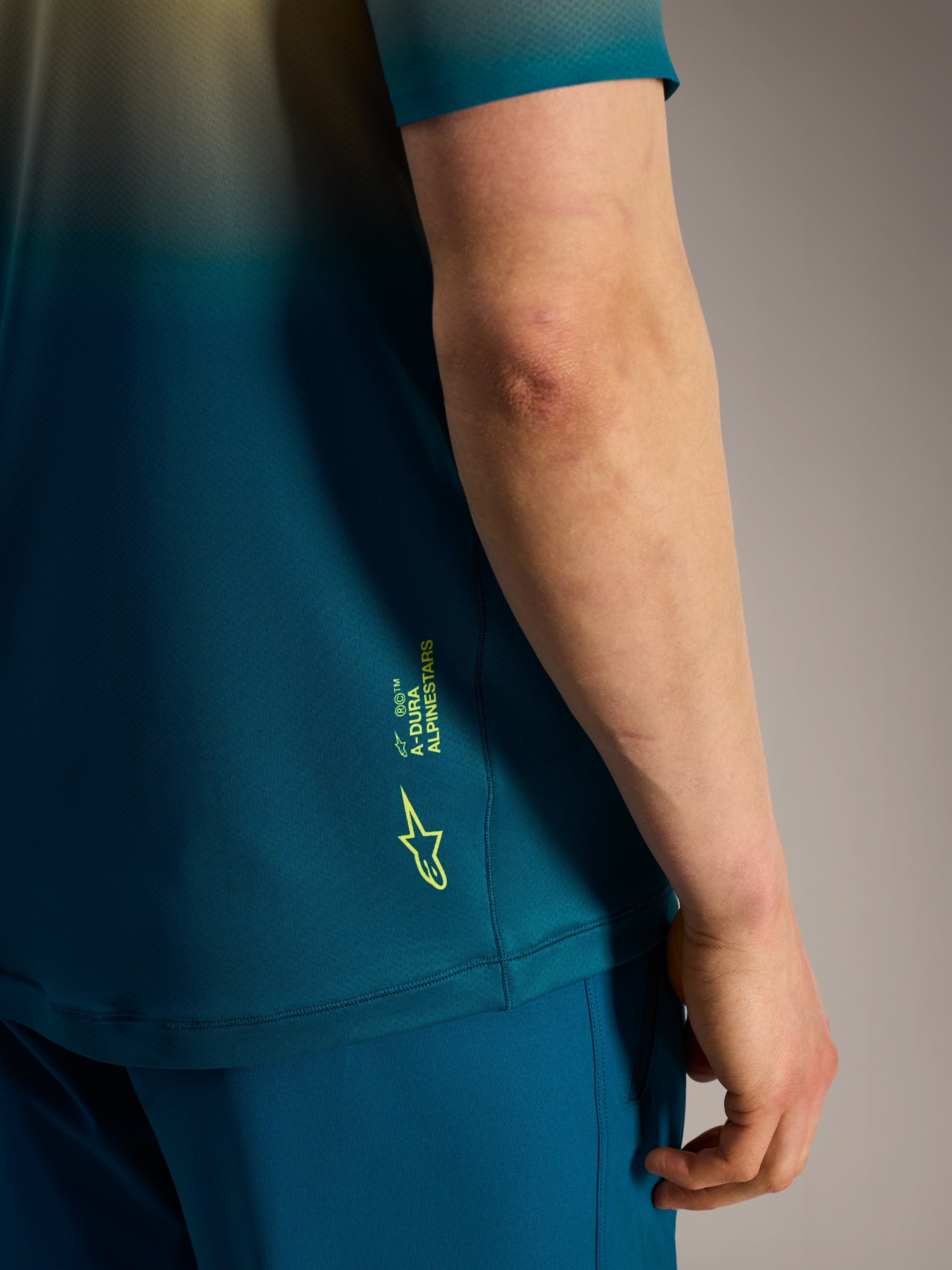 Détail du maillot Alpinestars A-Dura ASTARS 2.0 bleu-vert, avec logo et marquage jaune fluo sur le côté.