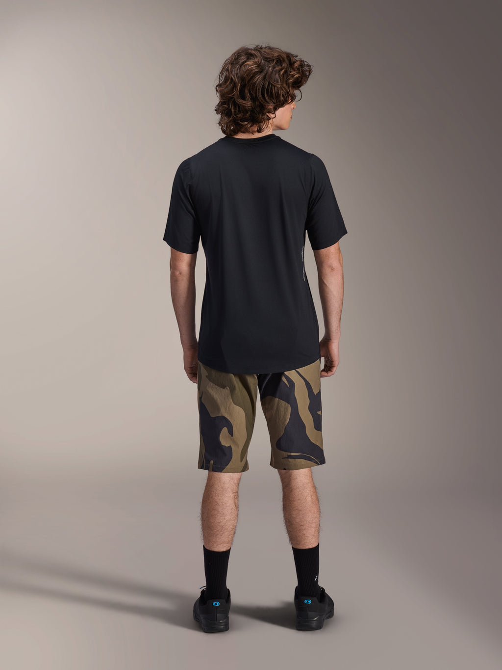 Maillot à manches courtes Alpinestars A-Dura Astar, noir, vue de dos sur un modèle avec un short de VTT à motif camouflage et des chaussures noires, vêtements techniques de VTT décontractés