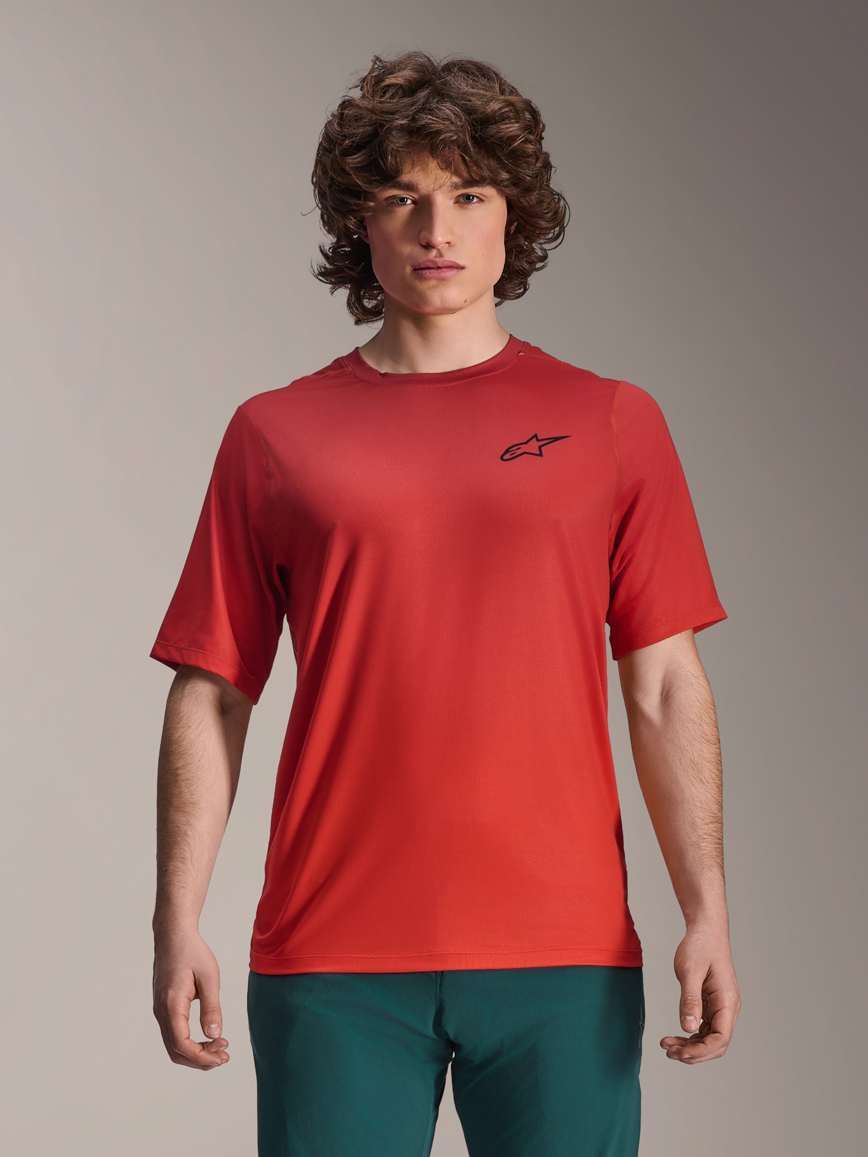 Alpinestars 2025 A-Dura Rise Jersey, Short Sleeve MTB Shirt, Spicy Orange, worn by a man, featuring a black Astars logo on the chest, paired with dark green pants, casual mountain biking performance apparel