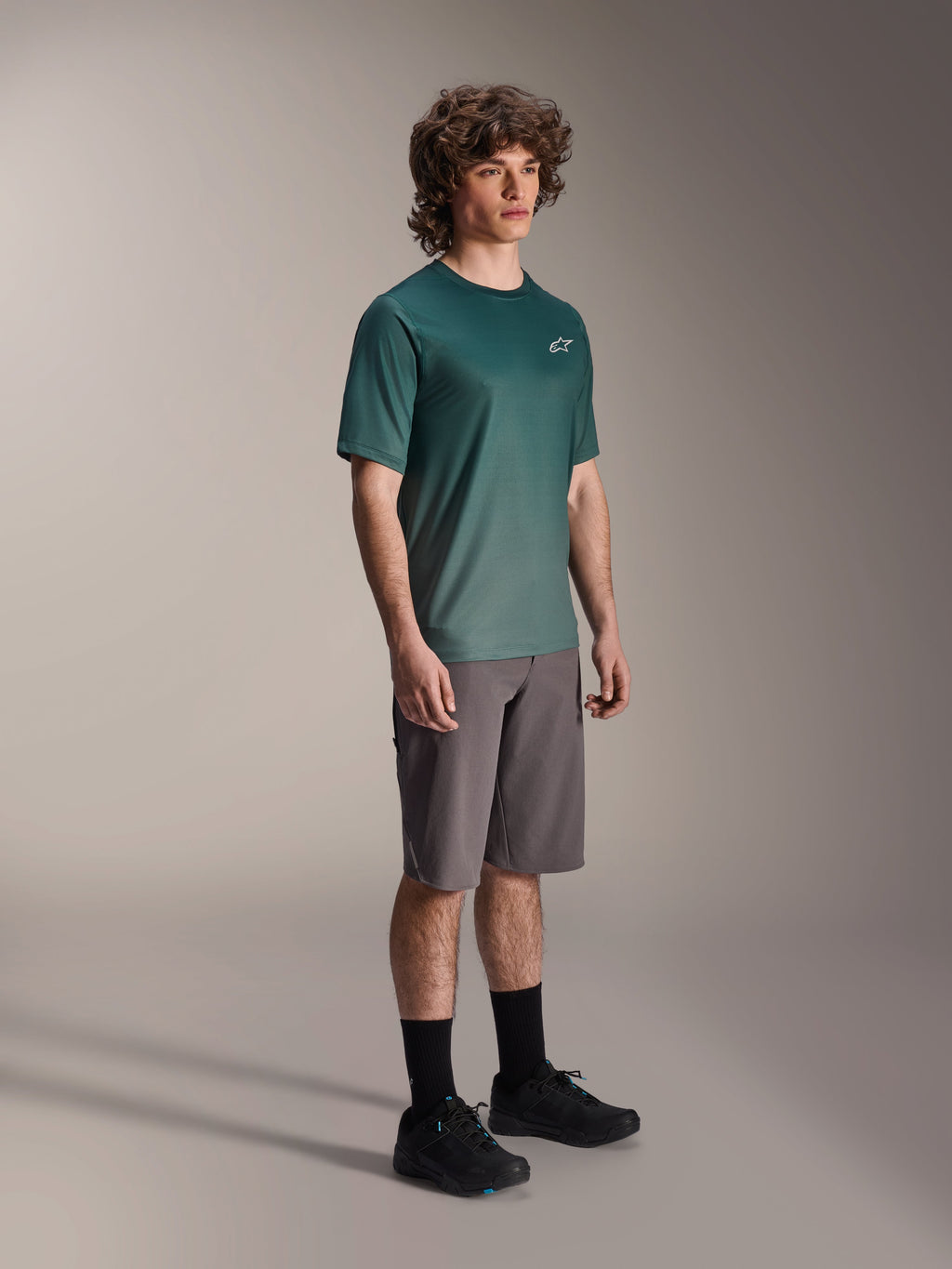 Alpinestars A-Dura Rise Jersey - Short Sleeve en Silver Pine, maillot de VTT vert porté par un mannequin avec un short gris et des baskets noires, tenue de VTT décontractée avec un petit logo blanc sur la poitrine