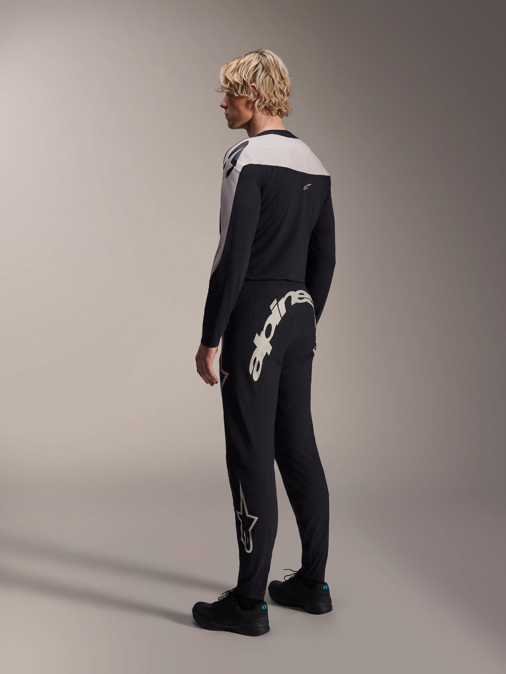 Maillot à manches longues Alpinestars A-Supra Melt, noir avec empiècements blancs aux épaules, vu de dos et de côté sur un modèle, associé à un pantalon de VTT noir avec un grand logo blanc, tenue de course VTT haute performance