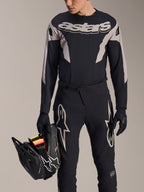 Ensemble de course VTT Alpinestars A-Supra Melt, maillot à manches longues et pantalon, noir et gris avec logos Astars blancs, modèle tenant un casque intégral noir et portant des gants assortis, équipement haute performance pour le VTT de descente et l'enduro