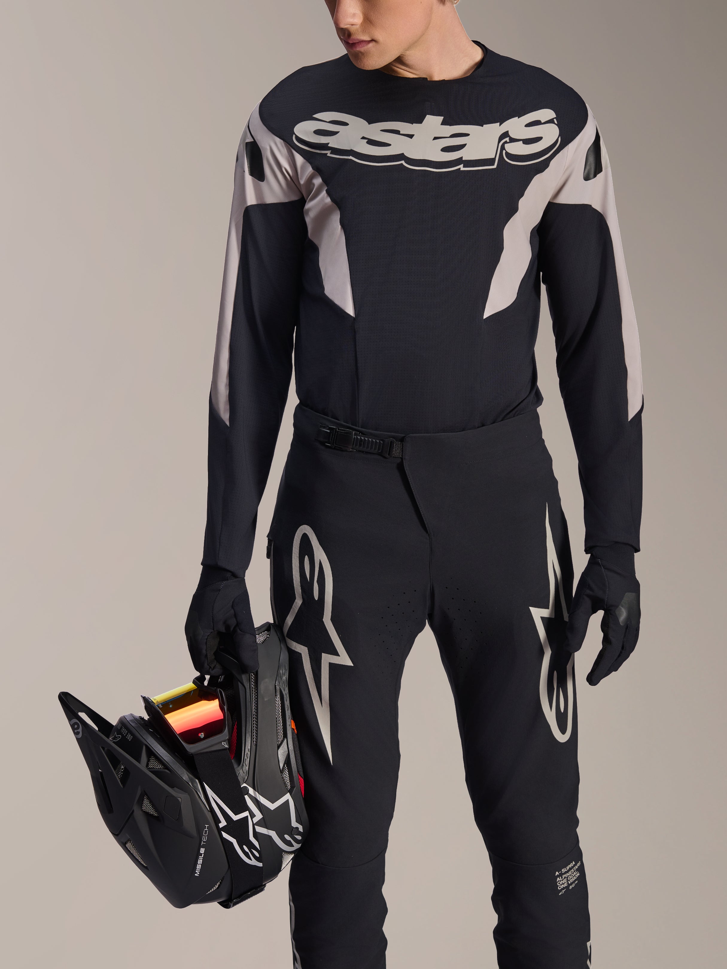 Ensemble de course VTT Alpinestars A-Supra Melt, maillot à manches longues et pantalon, noir et gris avec logos Astars blancs, modèle tenant un casque intégral noir et portant des gants assortis, équipement haute performance pour le VTT de descente et l'enduro