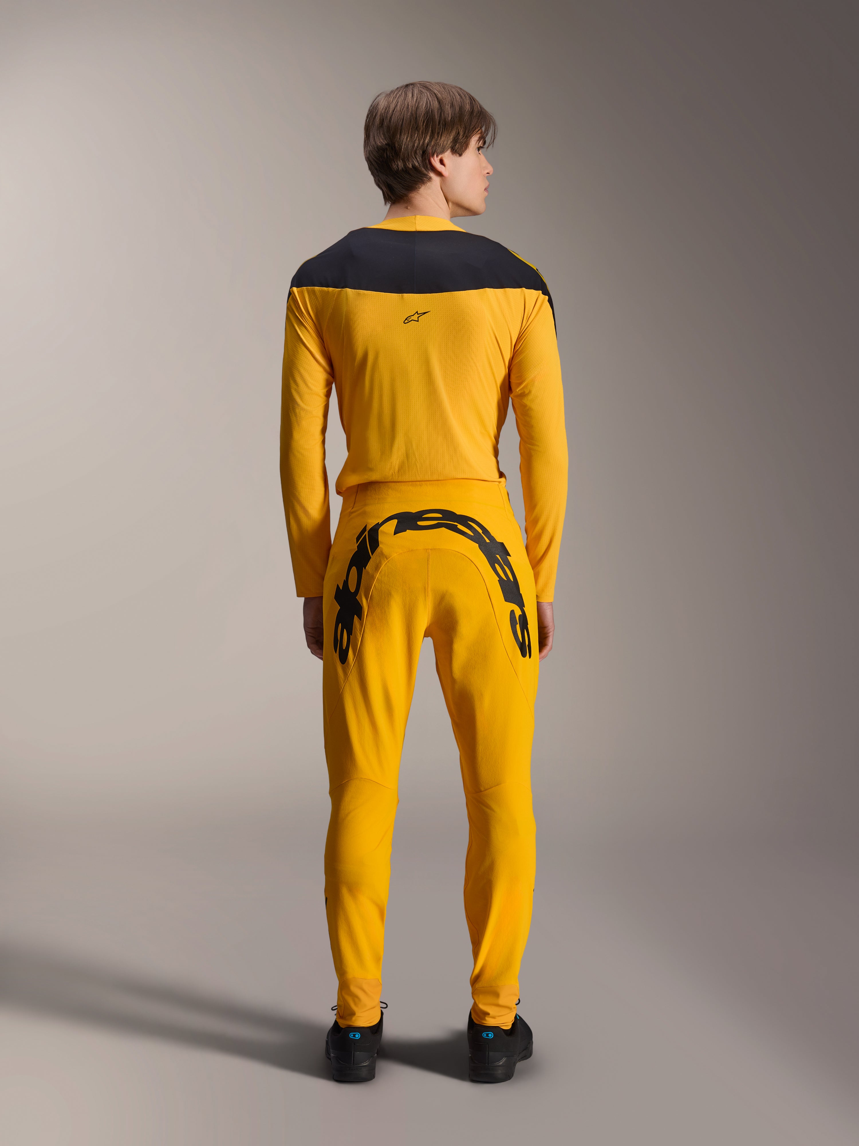 Alpinestars Limited Edition A-Supra Melt Gold Jersey, maillot de VTT à manches longues, jaune foncé, vue de dos, avec empiècements noirs aux épaules, porté avec un pantalon jaune assorti avec un large logo noir à l'arrière, équipement de cyclisme haute performance