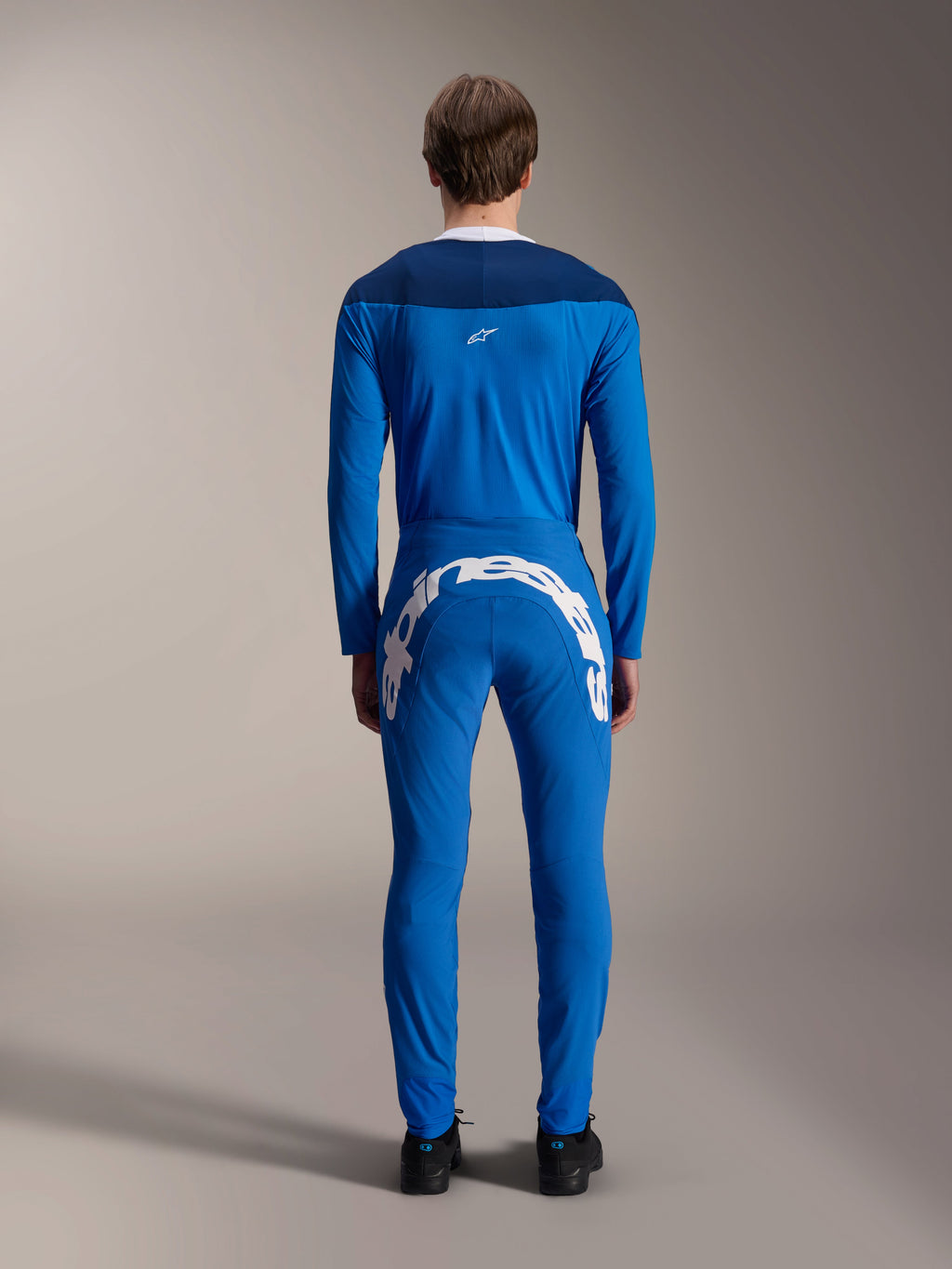 Pantalon Alpinestars A-Supra Melt, bleu, vue de dos avec un grand logo Alpinestars blanc sur l'empiècement arrière, porté avec un maillot à manches longues bleu assorti et des chaussures noires, équipement de course VTT haute performance