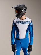 Modèle portant le maillot à manches longues Alpinestars A-Supra Melt bleu et blanc, avec le logo 'astars' sur la poitrine, associé à un pantalon de VTT bleu assorti, un casque intégral noir et un masque, tenue de course complète pour VTT.