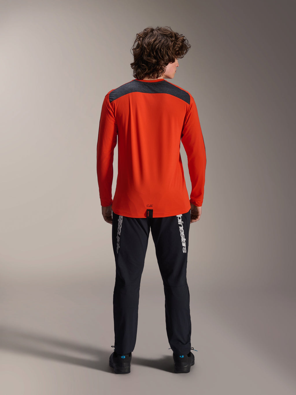 Maillot à manches longues Alpinestars A-Dura Insert Astar en Spicy Orange, vue de dos montrant l'empiècement gris anthracite aux épaules, porté avec un pantalon Alpinestars noir et des chaussures noires, photo de studio professionnelle pour équipement de VTT