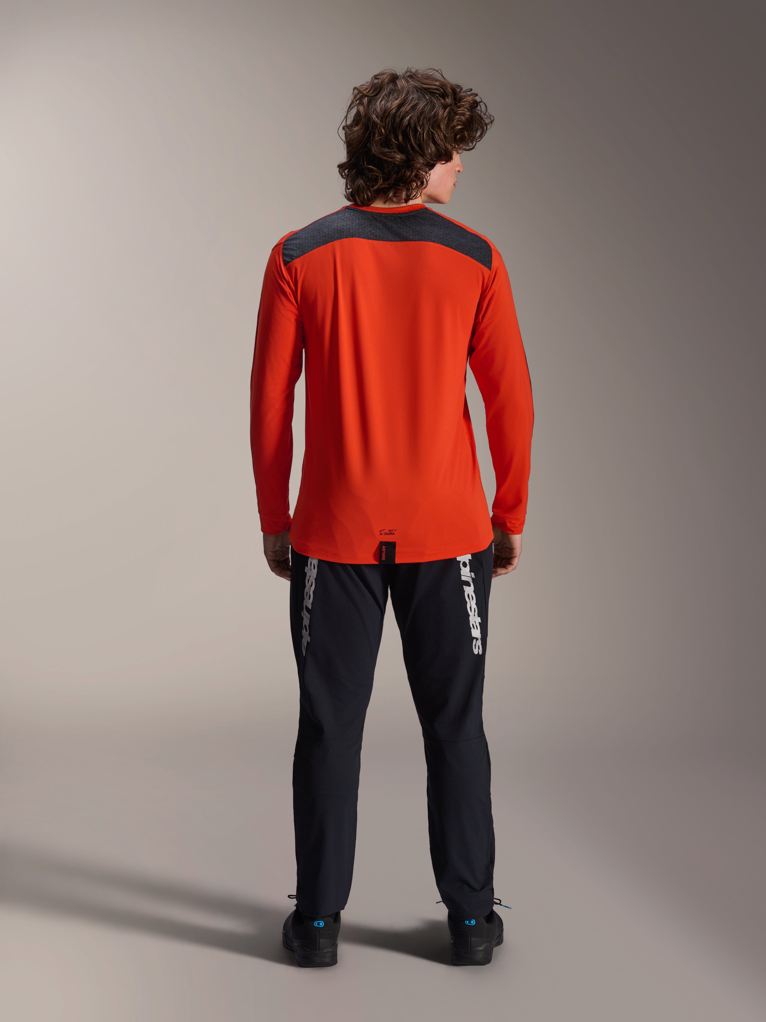 Maillot à manches longues Alpinestars A-Dura Insert Astar en Spicy Orange, vue de dos montrant l'empiècement gris anthracite aux épaules, porté avec un pantalon Alpinestars noir et des chaussures noires, photo de studio professionnelle pour équipement de VTT